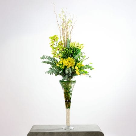 Tall floral arrangement with yellow lilies, green orchids, and eucalyptus in a clear glass vase with decorative white stones.