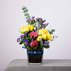 Bright yellow and purple flower arrangement with roses, statice, and eucalyptus in a BLUMEN square vase.