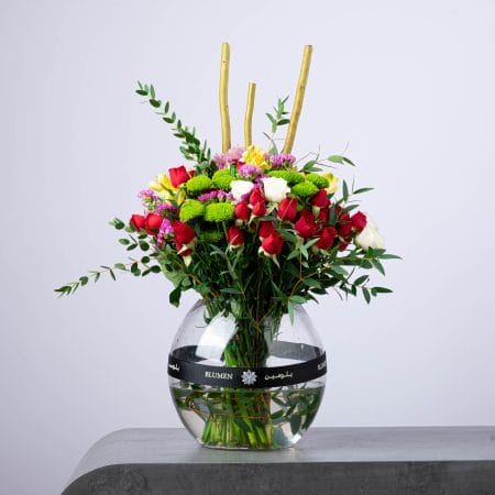 Colorful floral arrangement with red and white roses, green chrysanthemums, and bamboo accents in a round glass vase.