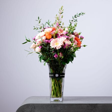 Elegant pastel flower arrangement with gerberas, roses, and eucalyptus in a tall clear BLUMEN vase.