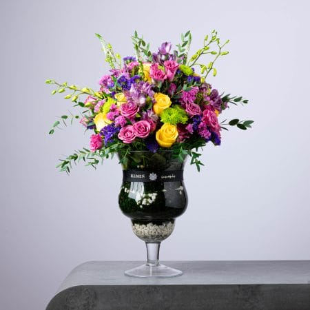 Vibrant floral arrangement with yellow and purple flowers in a tall glass vase with layered pebbles.