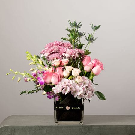 Soft pink floral arrangement with roses, chrysanthemums, orchids, and hydrangeas in a square glass vase.