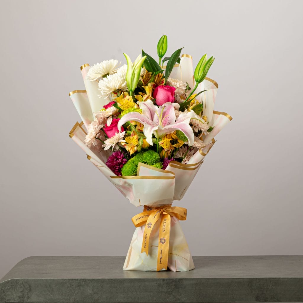 Vibrant bouquet of pink lilies, roses, and mixed flowers wrapped in white and gold paper with a yellow ribbon.