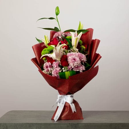 Elegant bouquet of pink lilies, red roses, and chrysanthemums wrapped in red paper with a white ribbon.