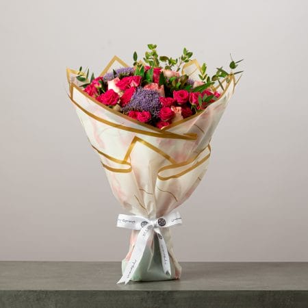 Romantic bouquet of pink and peach roses with purple statice flowers wrapped in white and gold paper with a white ribbon.