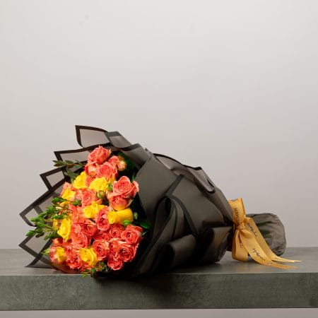 اقة زهور فاخرة من الورود البرتقالية والصفراء ملفوفة بورق أسود أنيق ومربوطة بشريط ذهبي يحمل شعار بلومن.