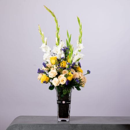 Tall yellow and white flower arrangement with gladiolus, roses, and statice in a clear BLUMEN vase.