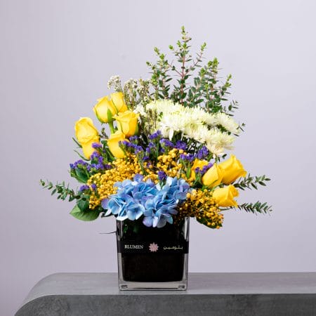 Colorful yellow, blue, and purple flower arrangement with hydrangeas and roses in a BLUMEN square vase.