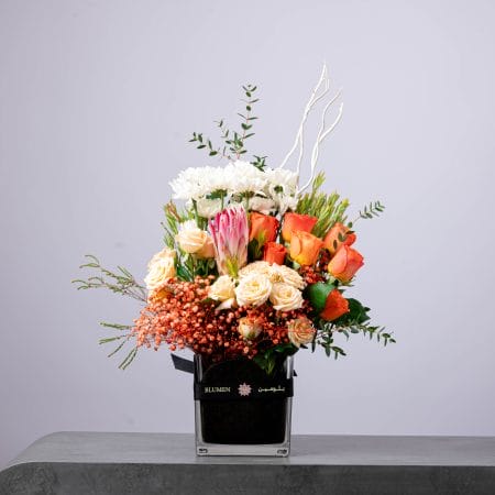 Vibrant floral arrangement with orange and cream roses, white chrysanthemums, and pink protea in a square glass vase.