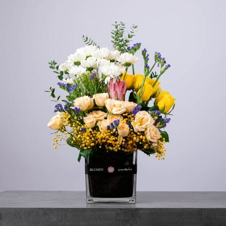 Floral arrangement with yellow roses, cream roses, and white chrysanthemums in a square vase.