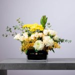 Elegant yellow and white floral arrangement in a rectangular BLUMEN glass vase.