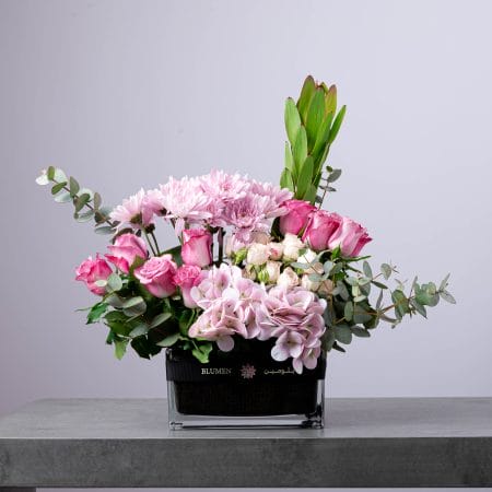 Colorful mixed flower arrangement with pink, yellow, and purple blooms in a round glass vase.
