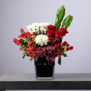 Elegant floral arrangement with red roses, white chrysanthemums, and burgundy hydrangeas in a black glass vase.