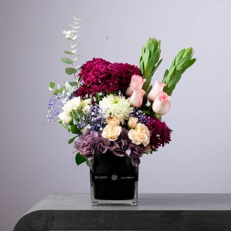 Elegant floral arrangement with pink roses, burgundy chrysanthemums, and white blooms in a black glass vase.