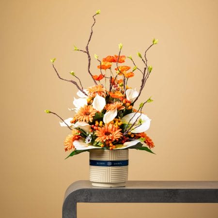 Artificial orange and white floral arrangement with calla lilies and gerberas in a cream geometric vase.