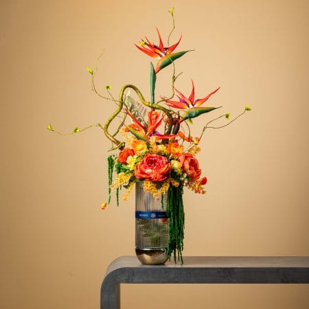 Artificial tropical arrangement with orange and red flowers in a tall ribbed glass vase.