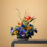 Artificial tropical floral arrangement with Birds of Paradise and blue flowers in a patterned pot.