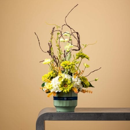 Artificial yellow and white floral arrangement with twigs in a mint-green pot.