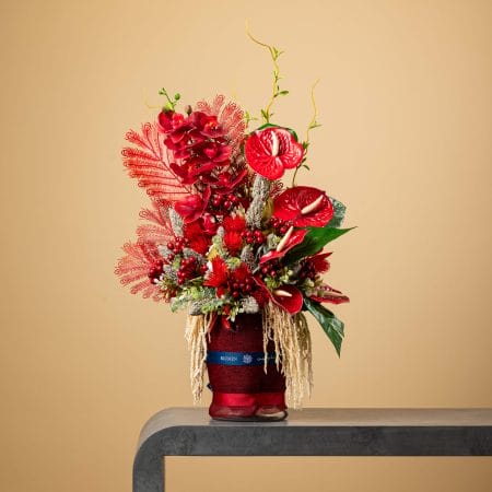 Artificial red floral arrangement with anthuriums, berries, and feathers in a red vase.