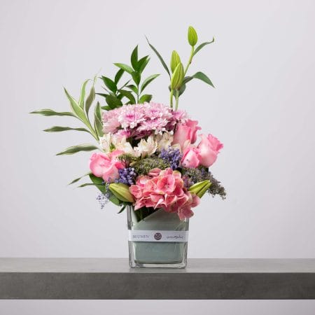 تنسيق زهور وردية وبيضاء أنيق يضم الورود والزنابق والكوبية في فازة زجاجية بشريط بلومن