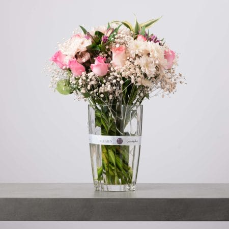 تنسيق زهور وردية وبيضاء ناعمة يضم الورود والأقحوان وورد الجبسوفيلا في فازة زجاجية طويلة من بلومن