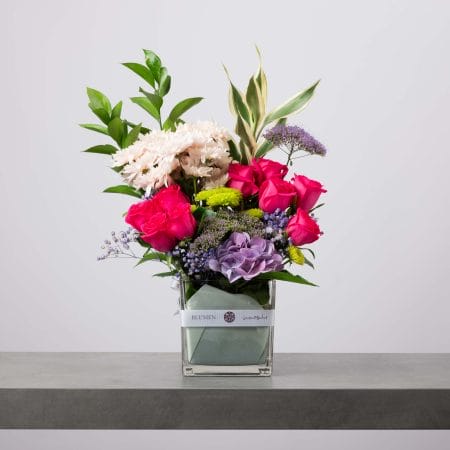 تنسيق زهور نابض بالحياة يضم ورود فوشيا وكوبية بنفسجية وزهور خضراء وأقحوان أبيض في فازة زجاجية شفافة من بلومن