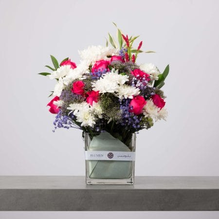 تنسيق زهور أنيق يضم ورود فوشيا وأقحوان أبيض وزهور بنفسجية صغيرة في فازة مربعة شفافة من بلومن.