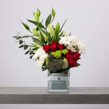 تنسيق زهور فاخر يضم ورودًا حمراء وأقحوانًا أبيض وأزهارًا خضراء وزنابق داخل فازة مربعة شفافة من بلومن.