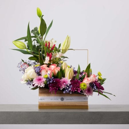 تنسيق زهور عصري يضم ورودًا وردية وزنابق بيضاء وأقحوانات بنفسجية ولمسات خضراء داخل صندوق مستطيل من بلومن مزين بمقبض ذهبي.