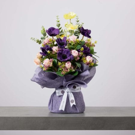 باقة زهور أنيقة تضم شقائق النعمان البنفسجية العميقة وورودًا وردية فاتحة وزهورًا صفراء ناعمة مغلفة بقماش بنفسجي مزين بشريط بلومن.