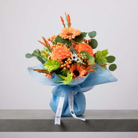باقة زهور مفعمة بالحيوية تضم جربيرا برتقالية وداليا وأوراقًا خضراء ولمسات بيضاء، مغلفة بقماش أزرق فاتح ومزينة بشريط بلومن.