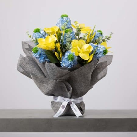 باقة زهور أنيقة تضم توليبات صفراء فاتحة وهدرانجيا زرقاء فاتحة وأوراقًا خضراء، مغلفة بقماش رمادي أنيق ومزينة بشريط بلومن أبيض.