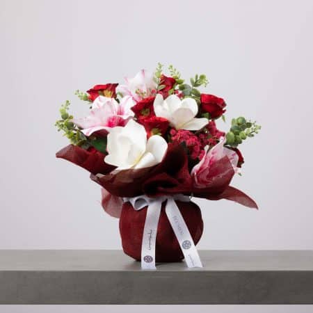 Luxurious bouquet with red roses, white magnolias, pink lilies, and eucalyptus, wrapped in deep burgundy fabric with a white BLUMEN ribbon.