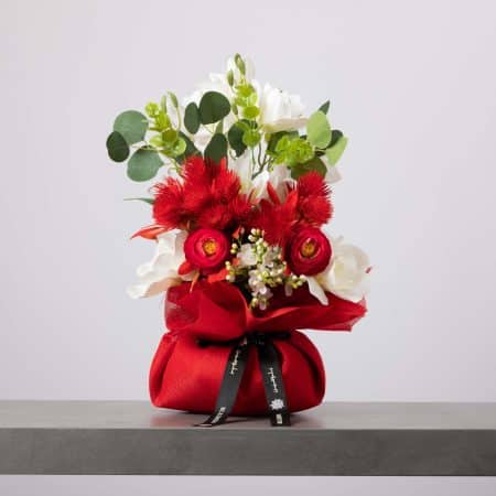 باقة نابضة بالحياة تضم زهور الحوذان الحمراء وزهورًا بيضاء غريبة وأوراق الأوكالبتوس، مغلفة بقماش أحمر لامع ومزينة بشريط بلومن أسود.