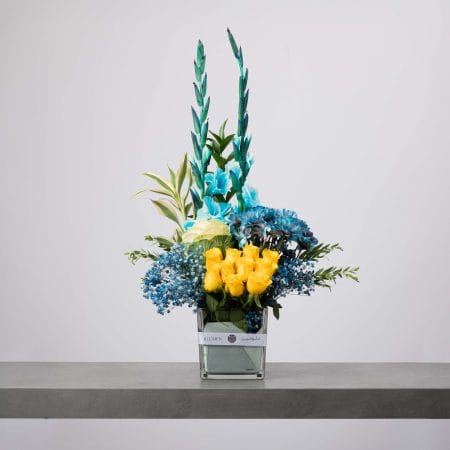 مزهرية زجاجية مربعة تحتوي على ورود صفراء زاهية وزنابق وسوسن أزرق وأقحوان أزرق، مزينة بأوراق خضراء ومرتبة بتصميم عمودي عصري على طاولة رمادية.