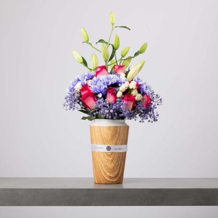 مزهرية خشبية طويلة بحافة بيضاء تحتوي على ورود وردية وأقحوان بنفسجي وزنابق فاتحة اللون، محاطة بزهور الجيبسوفيلا البنفسجية، مرتبة بأناقة على طاولة رمادية وخلفية ناعمة.