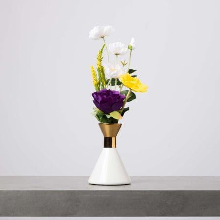 مزهرية أنيقة باللونين الأبيض والذهبي تحتوي على تنسيق بسيط من الزهور البيضاء والصفراء والبنفسجية الداكنة، موضوعة على طاولة رمادية وخلفية فاتحة.