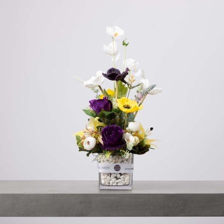 مزهرية زجاجية مربعة تحتوي على زهور بنفسجية وبيضاء وصفراء مرتبة بشكل عمودي مع أوراق خضراء وحصى بيضاء في القاع، موضوعة على طاولة رمادية وخلفية فاتحة.