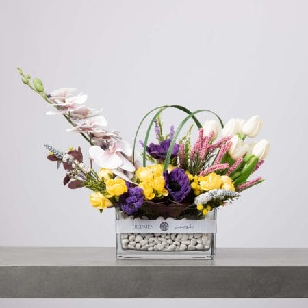 مزهرية زجاجية مستطيلة تحتوي على زهور الأوركيد البيضاء وورود صفراء وأنايمون بنفسجية وتوليب بيضاء مرتبة بشكل غير متماثل مع لمسات وردية وحصى بيضاء في القاع، موضوعة على طاولة رمادية وخلفية فاتحة.