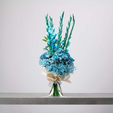 باقة طويلة من الزهور الزرقاء الزاهية تشمل الجلاديولس والأقحوان وزهور الجيبسوفيلا، ملفوفة بورق كريمي أنيق ومربوطة بشريط أبيض، موضوعة على طاولة رمادية وخلفية فاتحة.