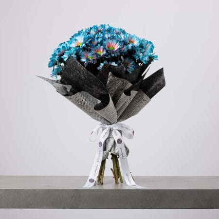 باقة من أزهار الأقحوان الزرقاء ملفوفة بورق أسود وفضي أنيق الملمس، ومربوطة بشريط أبيض، موضوعة على طاولة رمادية وخلفية فاتحة.