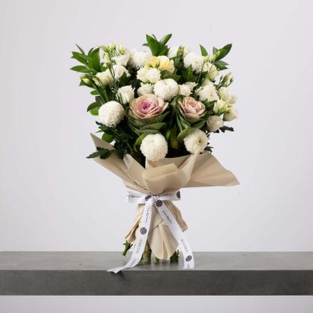 باقة من الزهور البيضاء والوردية الفاتحة ملفوفة بورق بيج ومربوطة بشريط أبيض، تحتوي على زهور كروية بيضاء وورود وردية باهتة وأوراق خضراء كثيفة، موضوعة على طاولة رمادية وخلفية فاتحة.
