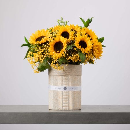 باقة مشرقة من زهور دوّار الشمس وزهور الميموزا الصفراء مرتبة في مزهرية خزفية بيج مزخرفة، موضوعة على سطح رمادي وخلفية فاتحة.