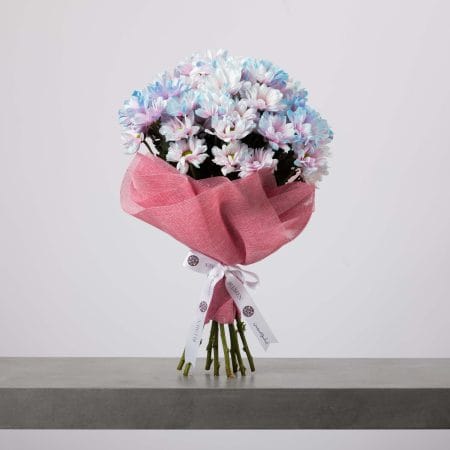 A pastel-colored bouquet of chrysanthemums in soft blue, lavender, and white hues, wrapped in pink mesh fabric and tied with a white ribbon, placed on a gray surface against a light background.