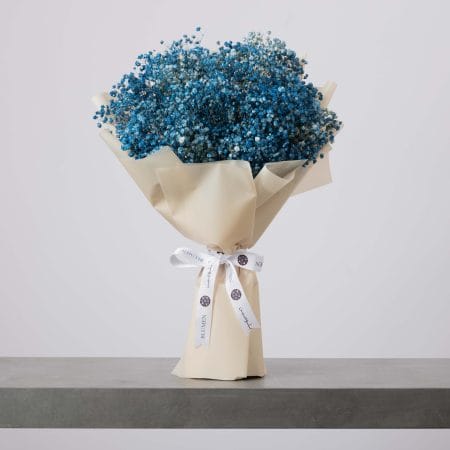 باقة من زهور التنفس الطفلي باللون الأزرق، ملفوفة بورق بيج ناعم ومربوطة بشريط أبيض، موضوعة على سطح رمادي وخلفية فاتحة.