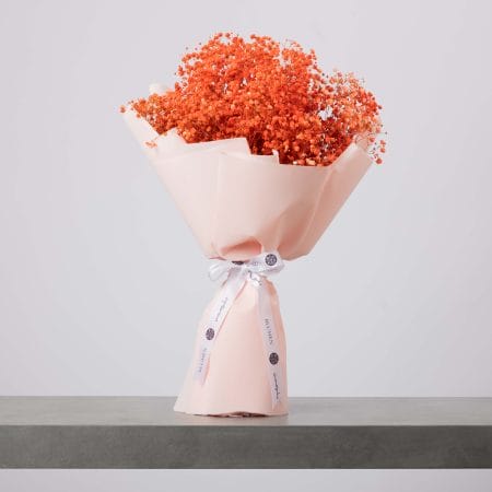 باقة من زهور التنفس الطفلي باللون البرتقالي المرجاني، ملفوفة بورق خوخي ناعم ومربوطة بشريط أبيض، موضوعة على سطح رمادي وخلفية فاتحة.