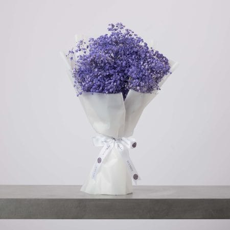 باقة من زهور التنفس الطفلي باللون البنفسجي الفاتح، ملفوفة بورق أبيض لامع ومربوطة بشريط أبيض، موضوعة على سطح رمادي وخلفية فاتحة.