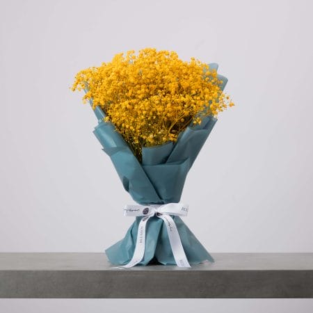 باقة من زهور التنفس الطفلي باللون الأصفر الزاهي، ملفوفة بورق أزرق مخضر ومربوطة بشريط أبيض، موضوعة على سطح رمادي وخلفية فاتحة.