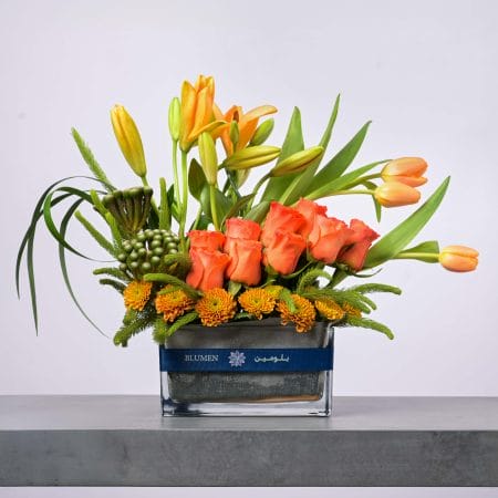 نسيق زهور أنيق يحتوي على ورود برتقالية وزنابق وتوليب صفراء في مزهرية زجاجية عصرية من بلومن.