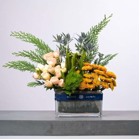 تنسيق زهور أنيق يضم ورود كريمية وذهبية وأوراق خضراء وزهور شوكية زرقاء في مزهرية زجاجية عصرية من بلومن.